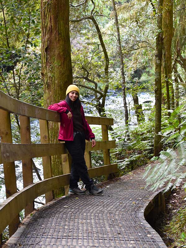 Camila en ruta por el bosque nativo Mujer sonriendo en un sendero entre árboles nativos en Nueva Zelanda.