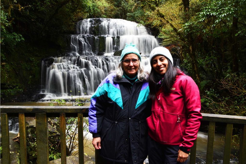 Amistad y naturaleza en Nueva Zelanda Dos mujeres sonrientes frente a una cascada natural.