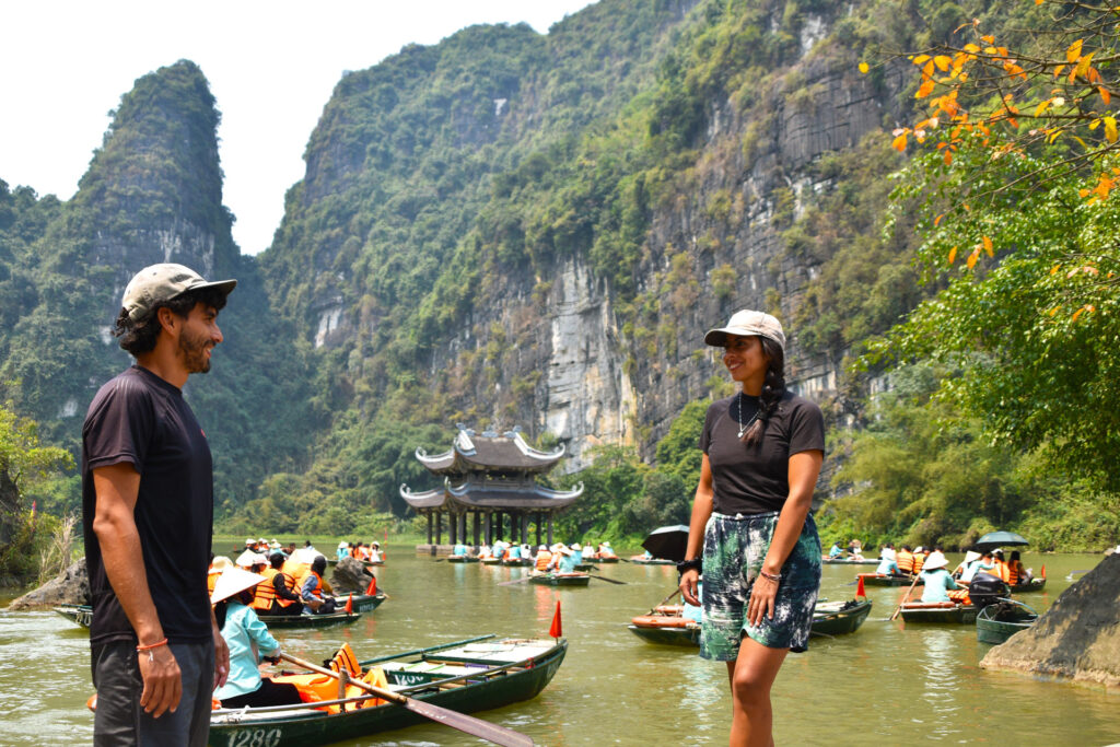 John y Camila conversando a la orilla del río con botes de remos al fondo en su Ruta en Ninh Binh Vietnam.