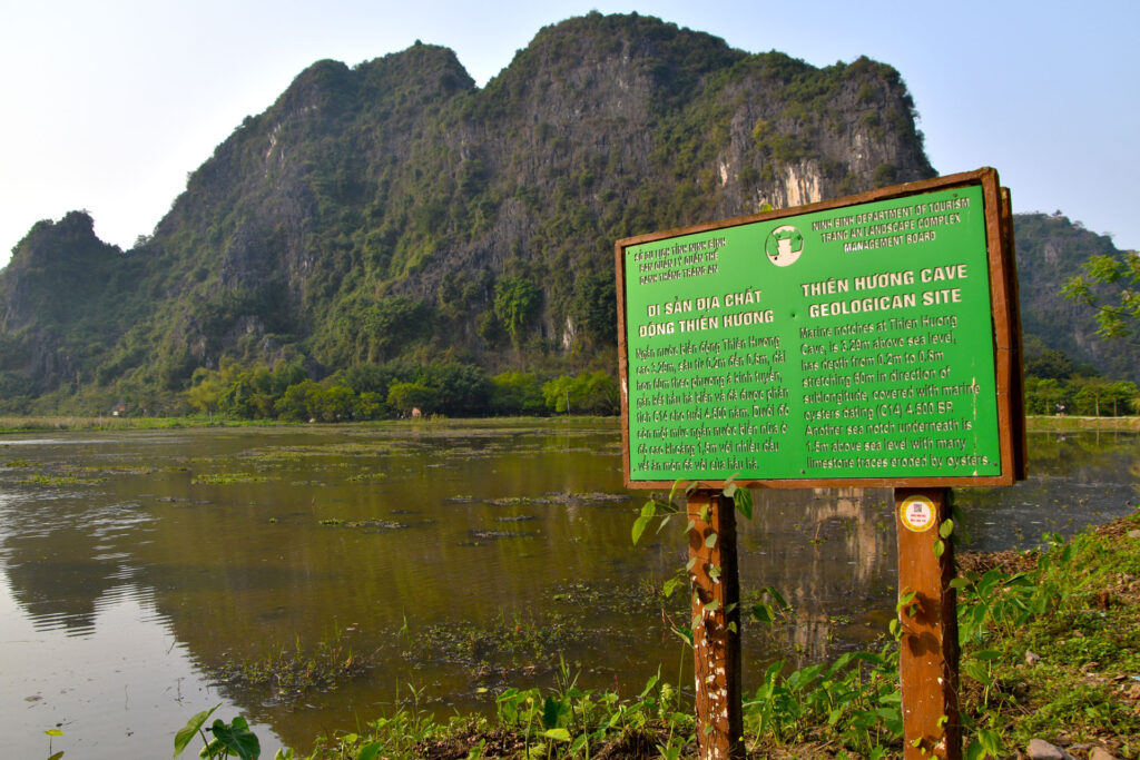 Cartel informativo de color verde situado frente a una zona de humedales y montañas en Vietnam.